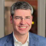 Headshot of Jay Scanlan in blue blazer and office buildings behind