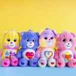 pink, blue and lavendar Care Bear stuffed animals sitting in front of yellow background