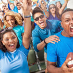 A large group of young adult football fans in outdoor stadium seats and dressed in blue clothing yell and cheer with excitement for their team.