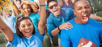 A large group of young adult football fans in outdoor stadium seats and dressed in blue clothing yell and cheer with excitement for their team.